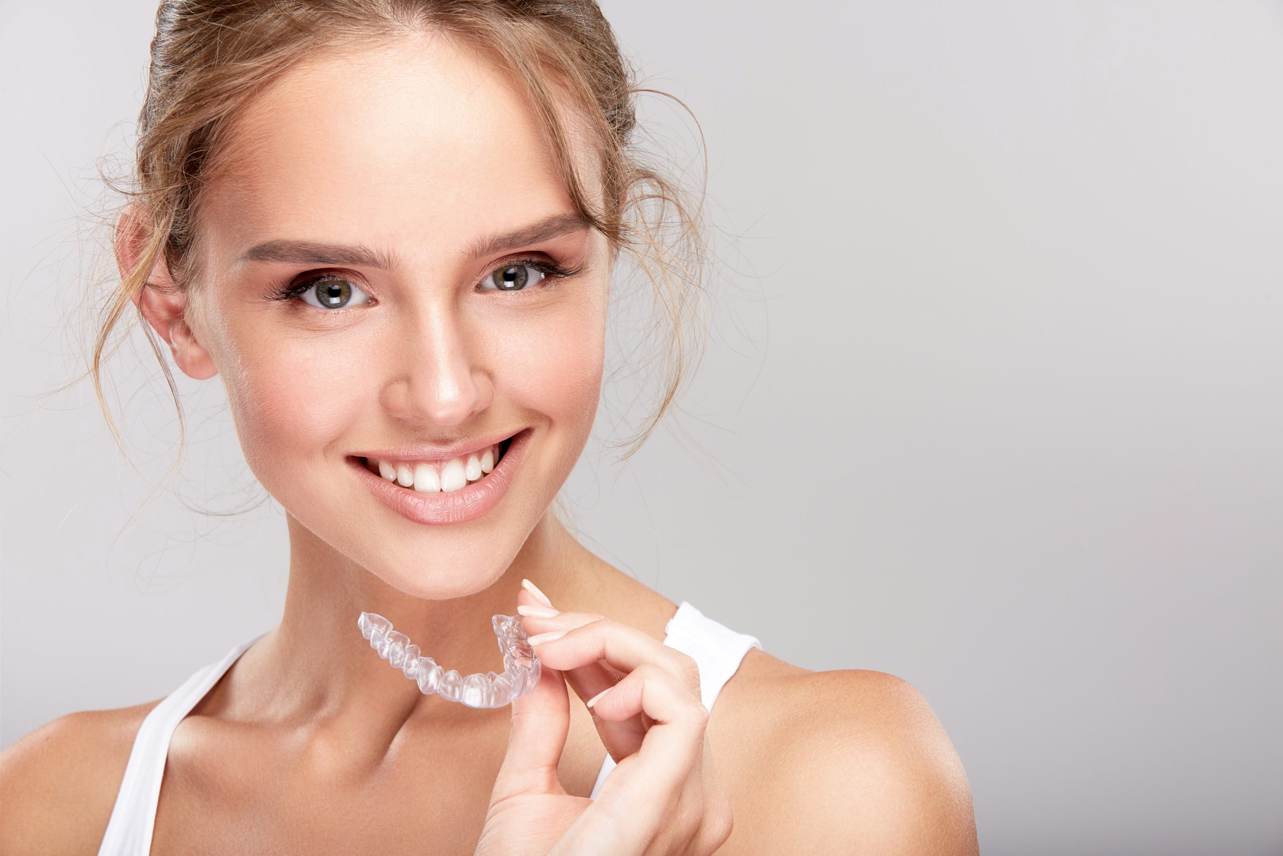 Young blonde-haired woman wearing white tank top and holding Invsialign - Is Invisalign Painful?